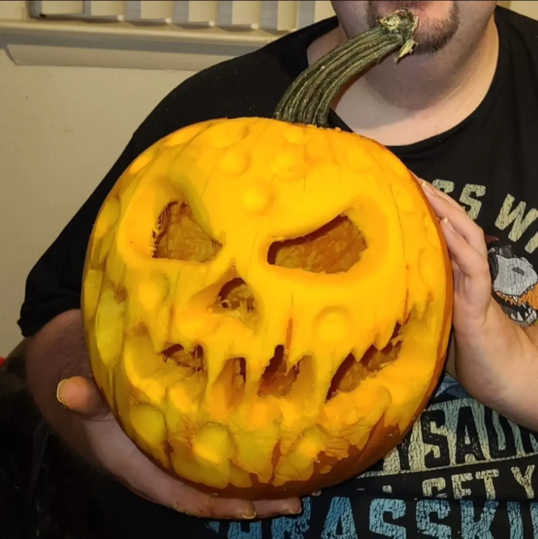 A person holds a large, intricately carved Halloween pumpkin with a menacing face, its stem stuck in their mouth. The…