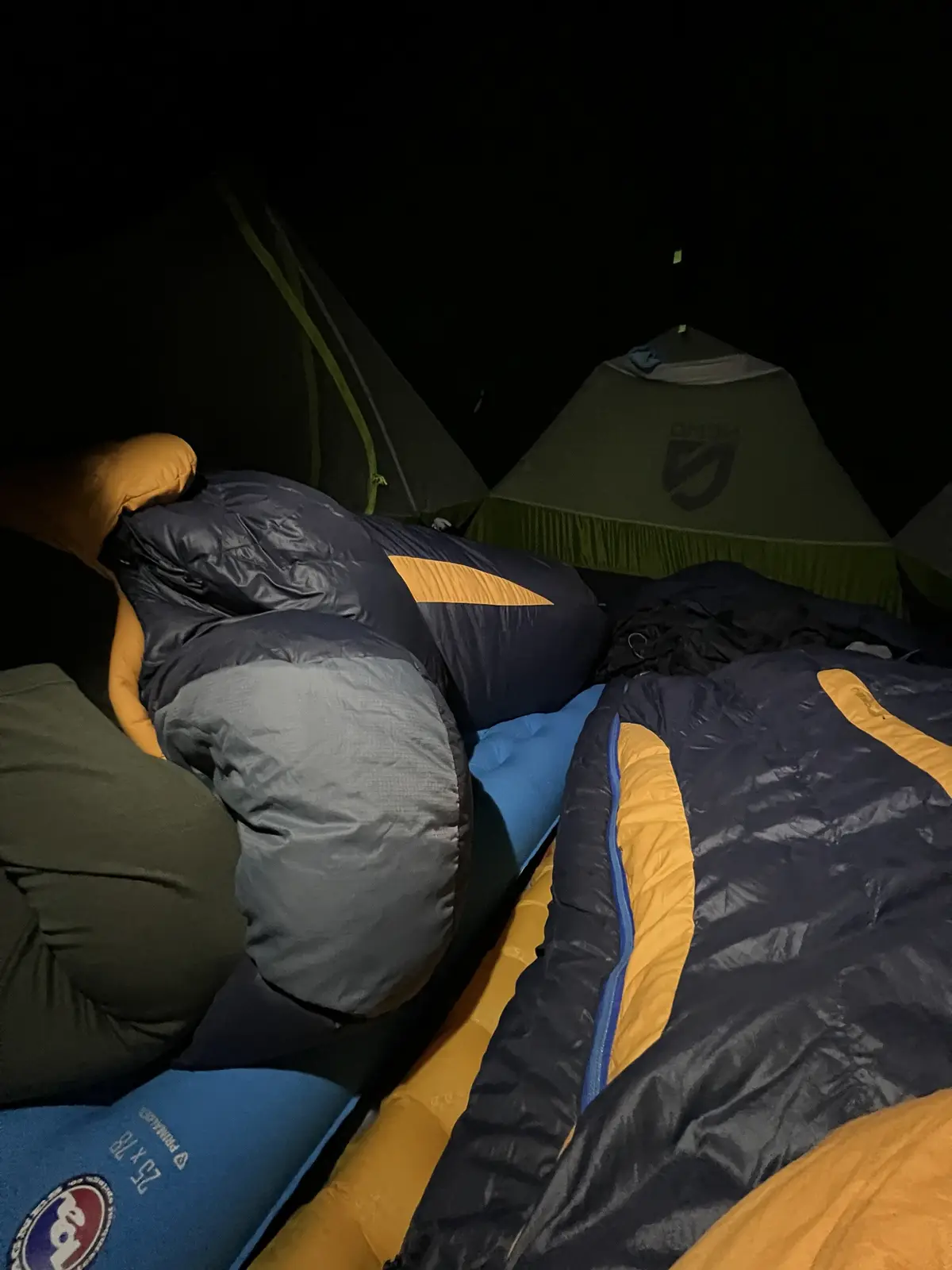 Inside a dark tent at night, two people are sleeping in sleeping bags, one with a blue and yellow sleeping bag and the other…