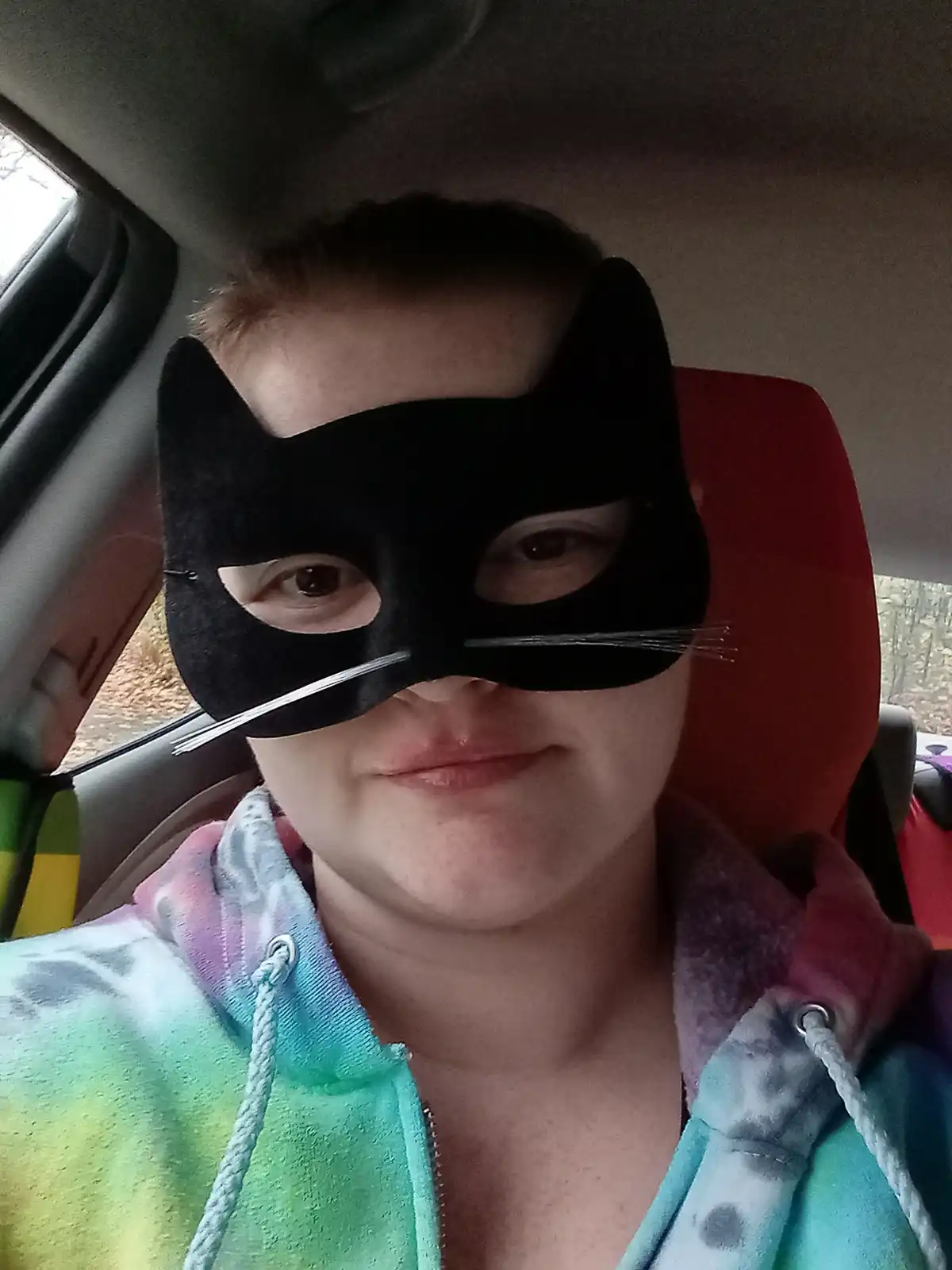 A person wearing a black cat mask with whiskers sits in a car, smiling slightly while wearing a colorful tie-dye hoodie. The…