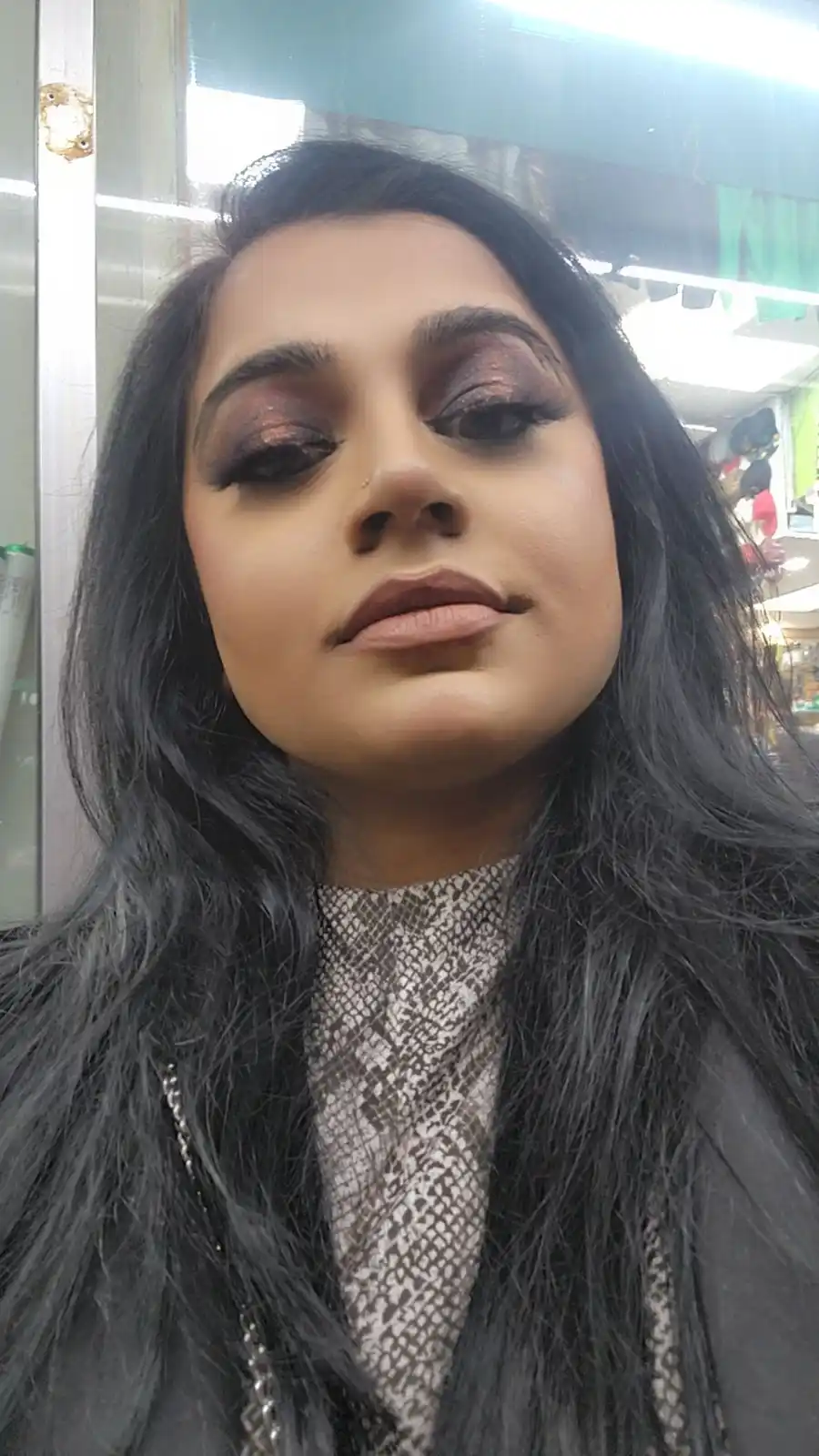 A selfie of a woman with long dark hair, heavy smoky eye makeup, and a neutral expression, wearing a snakeskin-patterned…