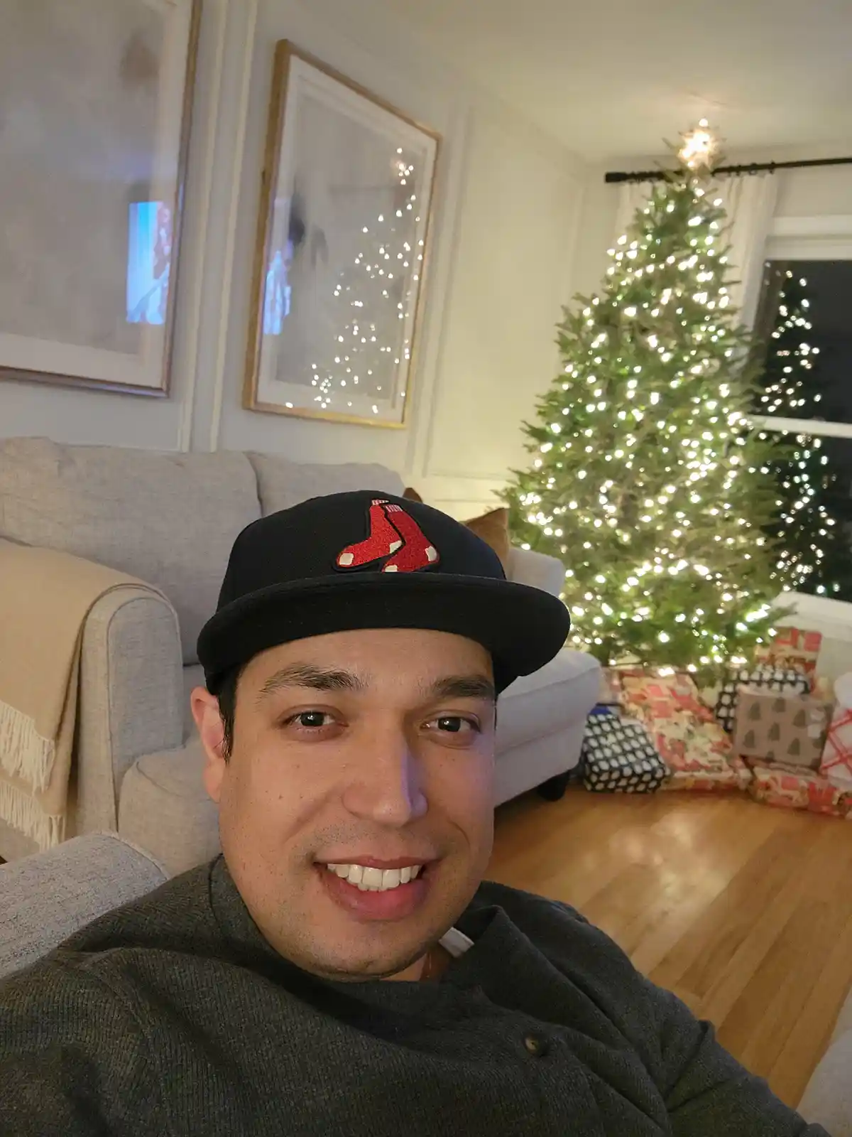 A man wearing a black Boston Red Sox baseball cap smiles at the camera while sitting on a couch in a living room decorated…