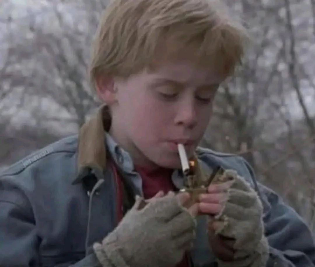 A young boy with blond hair, wearing a blue jacket and fuzzy gloves, is shown lighting a cigarette with a lighter. He is…