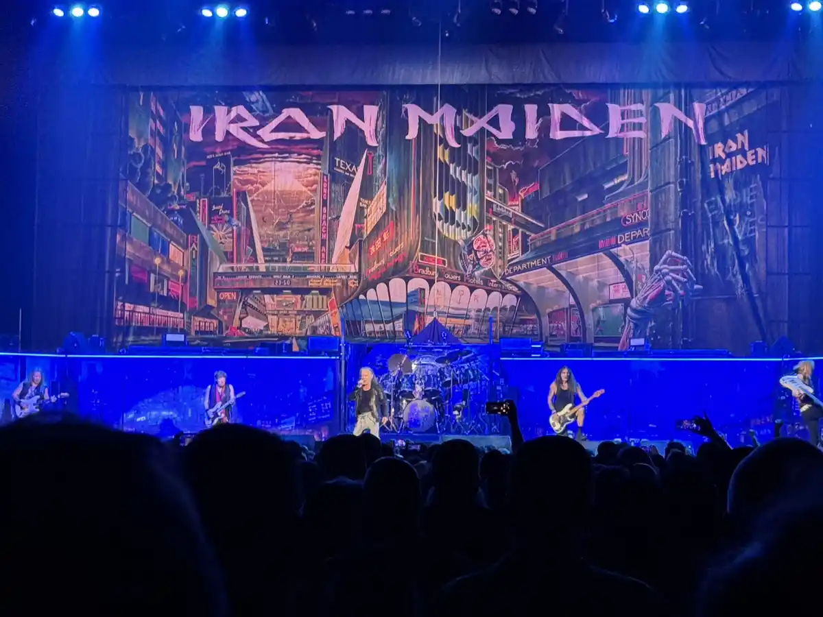 A live concert photo of the band Iron Maiden performing on stage with a large backdrop displaying their iconic 'The Number…