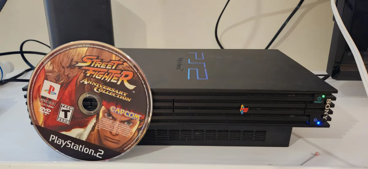 A black PlayStation 2 console sits on a white surface with a 'Street Fighter Anniversary Collection' game disc resting on…
