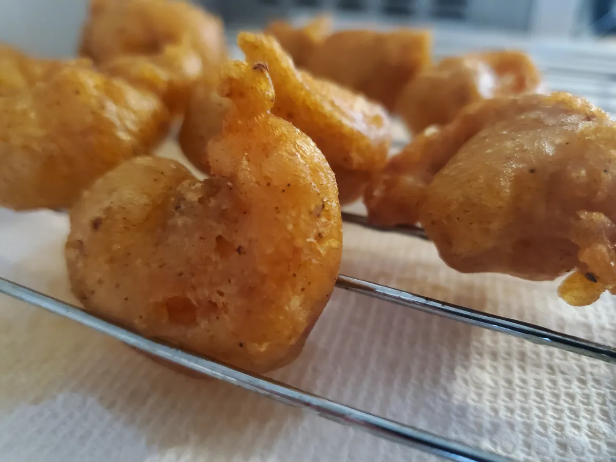 A close-up photo shows golden-brown, crispy fried shrimp resting on a metal rack over a white paper towel. One shrimp in the…