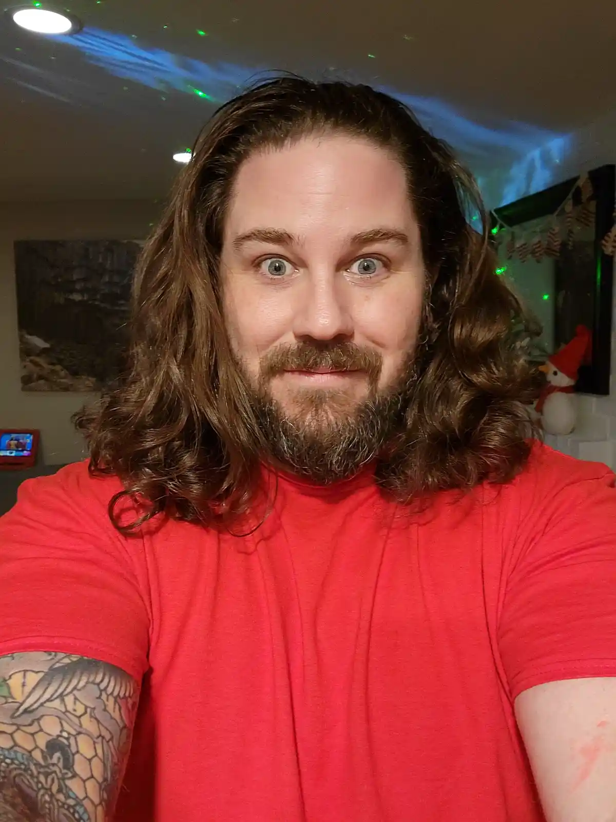 A man with long brown hair, a beard, and a red t-shirt takes a selfie with a surprised or exaggerated expression. He has a…