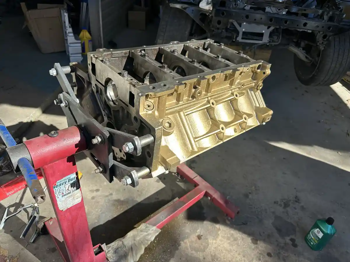 A freshly painted gold engine block is mounted on a red engine stand in a garage, with a partially disassembled vehicle…