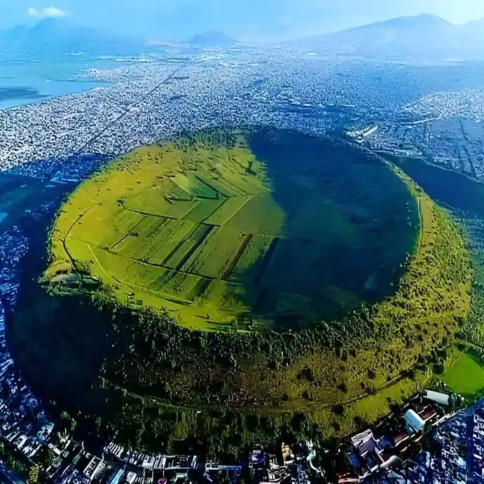 An aerial view shows a large, circular, green, crater-like structure surrounded by a dense urban area with buildings and…