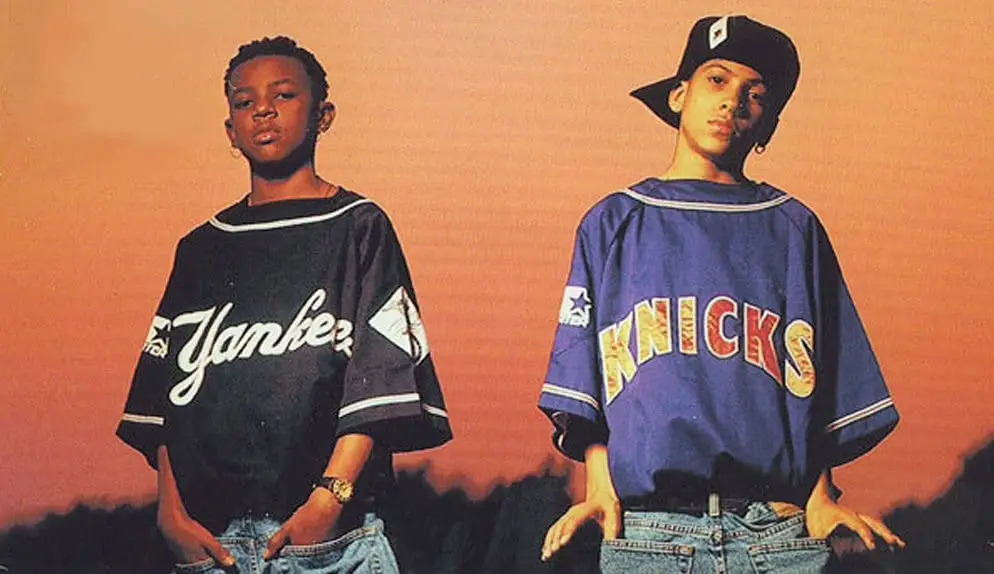Two young Black boys stand confidently against a sunset backdrop, one wearing a black oversized 'Yankees' baseball jersey…