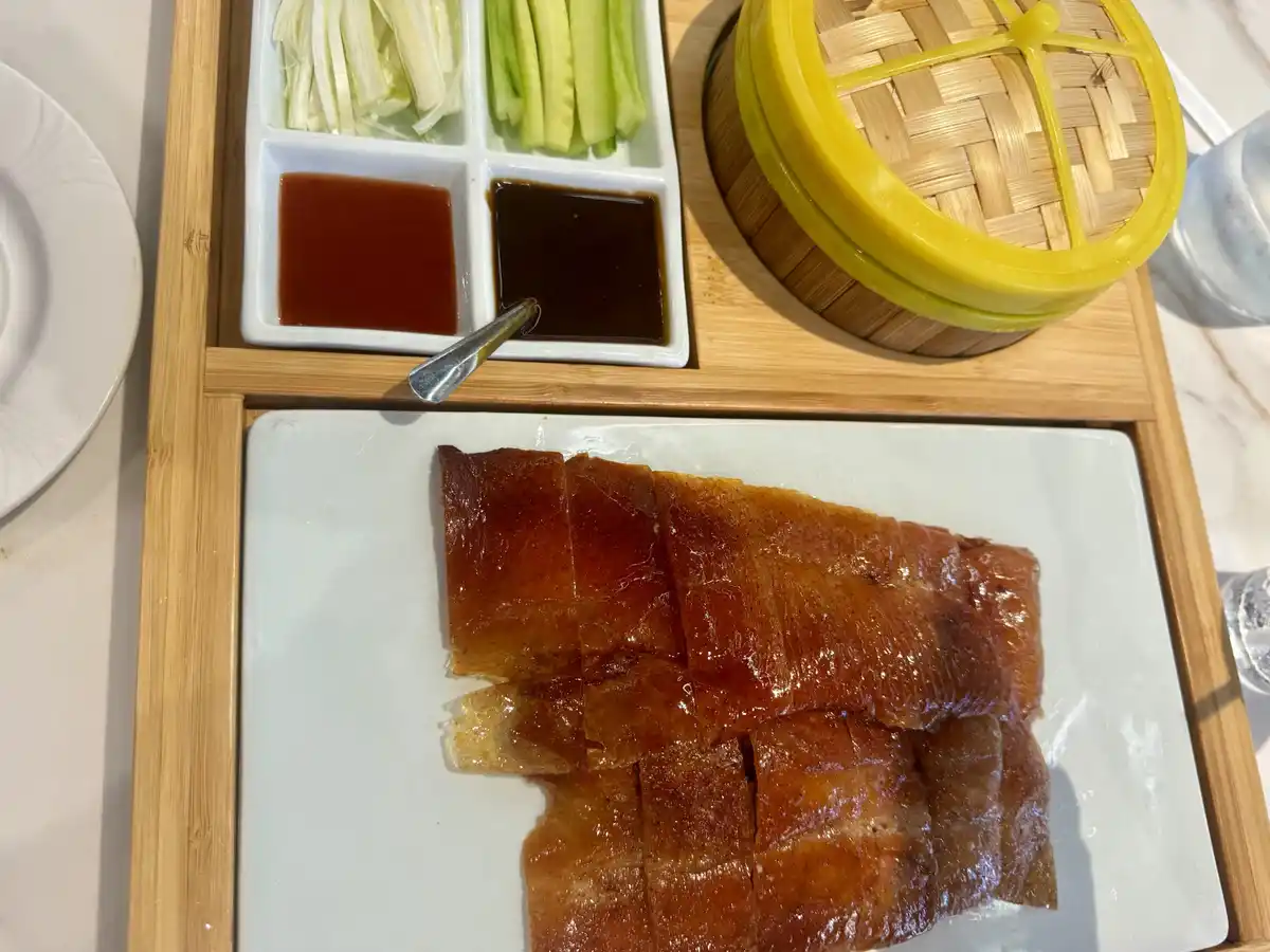 A close-up photo of a plate of sliced Peking duck with glossy, crispy skin, served with traditional accompaniments including…
