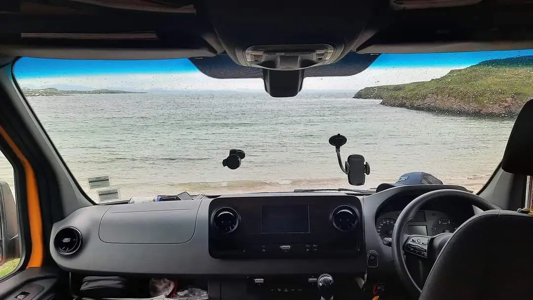 A first-person view from inside a van's driver's seat, looking out the windshield at a calm ocean with green hills on the…