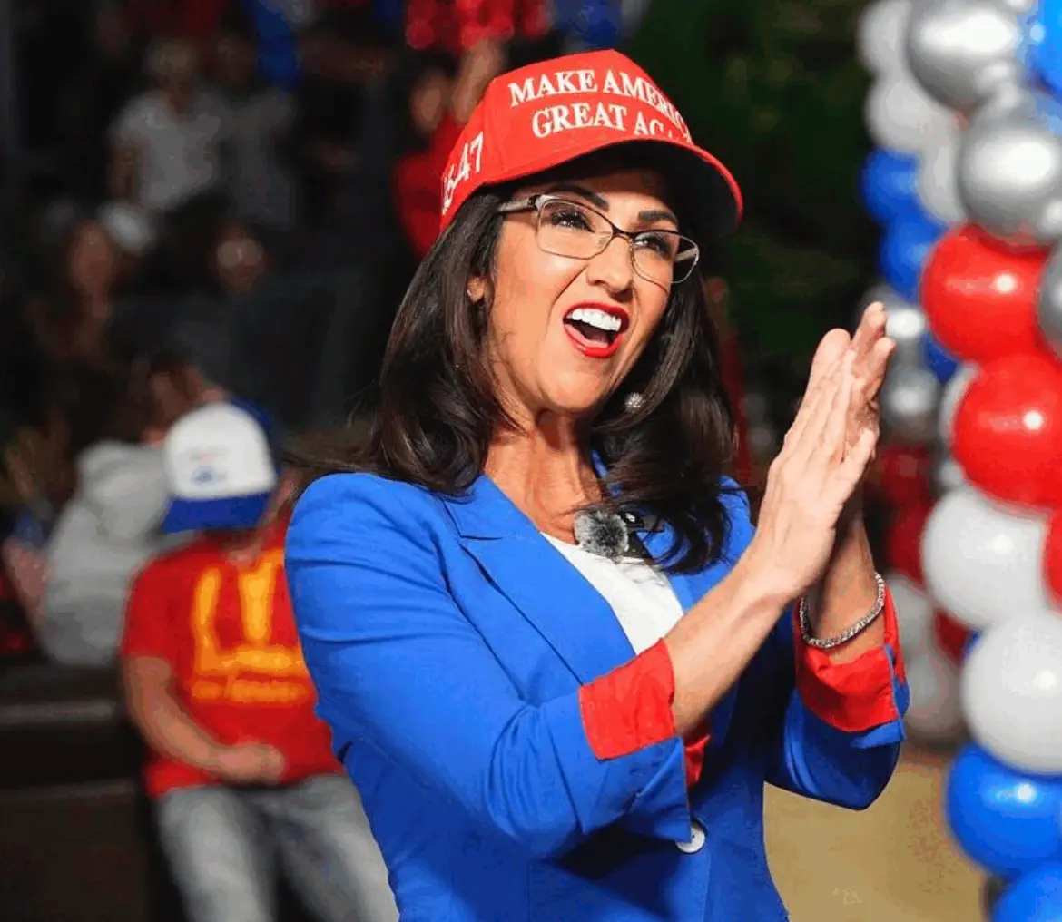 A woman with dark hair and glasses, wearing a blue blazer with red cuffs and a red 'Make America Great Again' cap, claps…