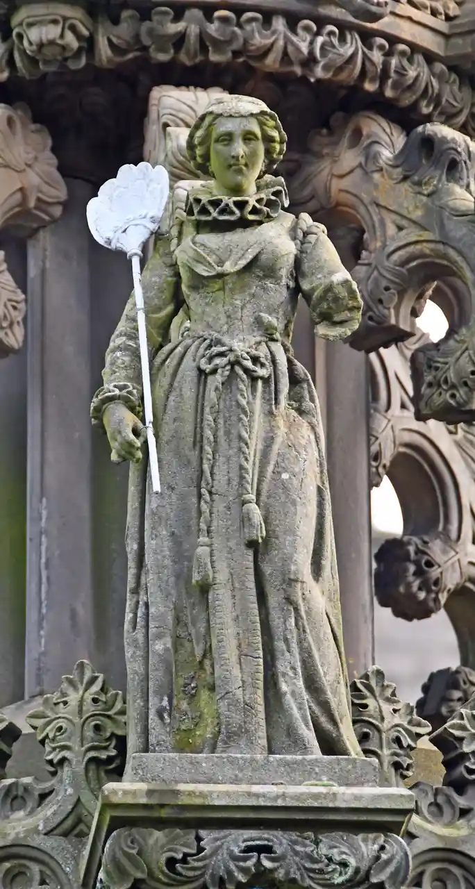A weathered stone statue of a woman in historical dress, holding a broom, stands on an ornate pedestal. The overlaid text…