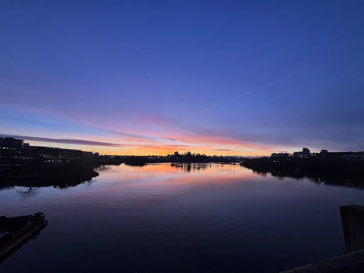 A serene twilight photo of a calm river reflecting a vibrant orange and pink sunset against a deep blue sky, with…