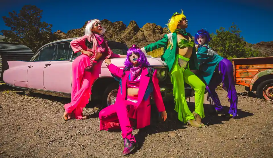 Four people in brightly colored, flamboyant outfits pose confidently around a pink vintage car in a desert setting. Each…