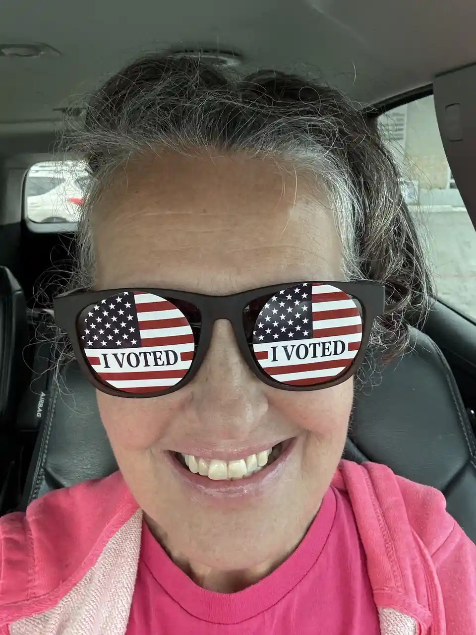 A smiling woman with gray hair sits in a car wearing black sunglasses with American flag lenses that each display the text…