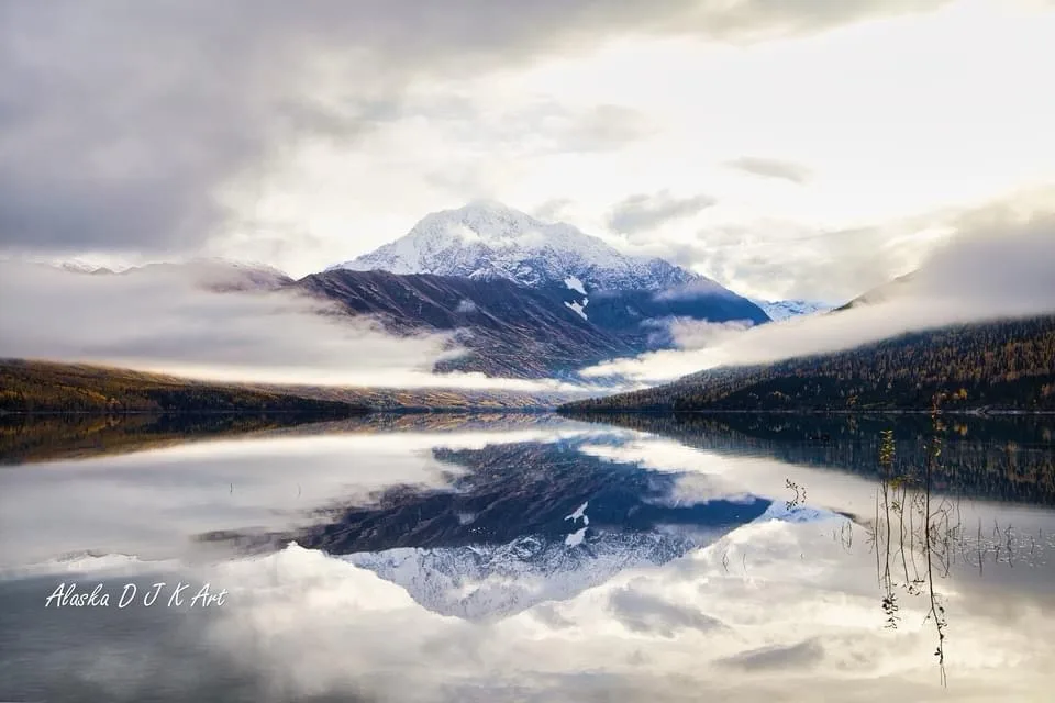 A serene landscape photograph shows a snow-capped mountain partially obscured by clouds, perfectly mirrored in a calm lake…
