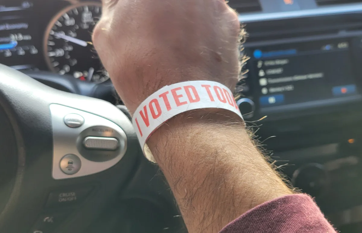 A close-up photo shows a person's hairy wrist inside a car, wearing a white wristband with red text that reads 'I VOTED…