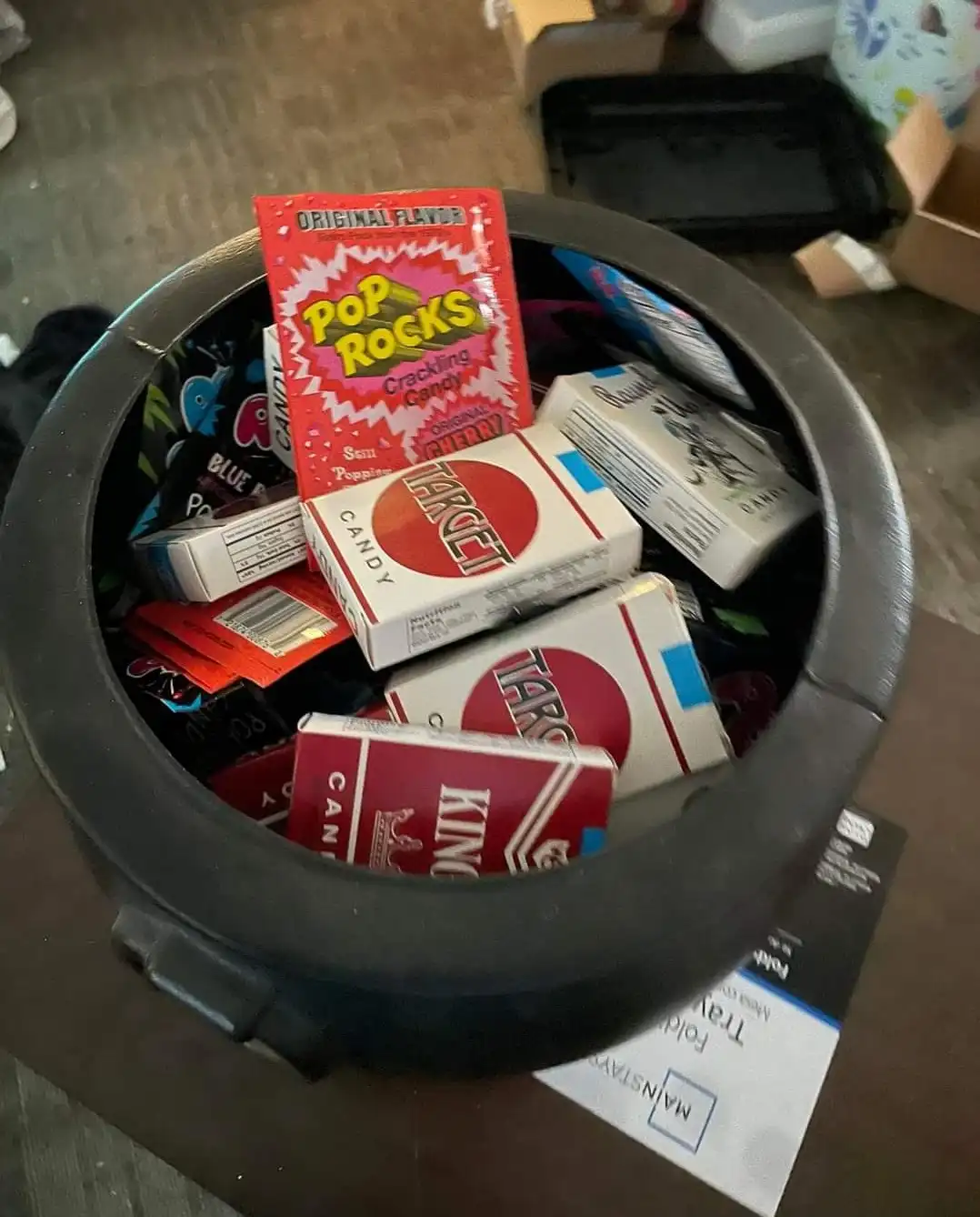 A black pressure cooker sits on a table filled with various candy boxes, including a prominent red box of Pop Rocks candy…
