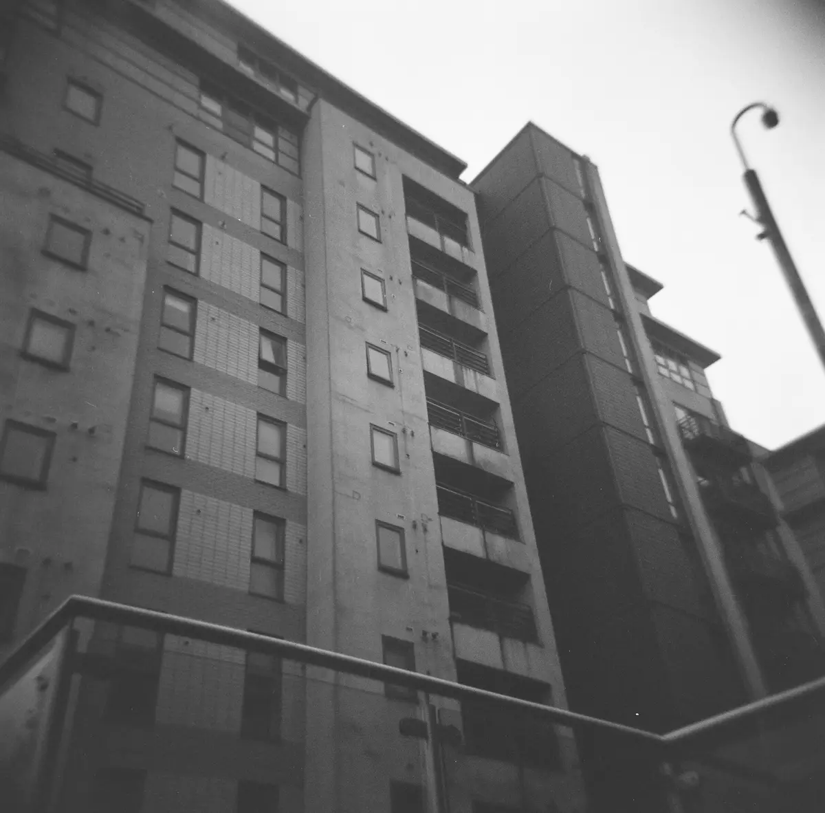A black-and-white, low-angle photograph of a tall, imposing concrete apartment building with many windows and balconies…