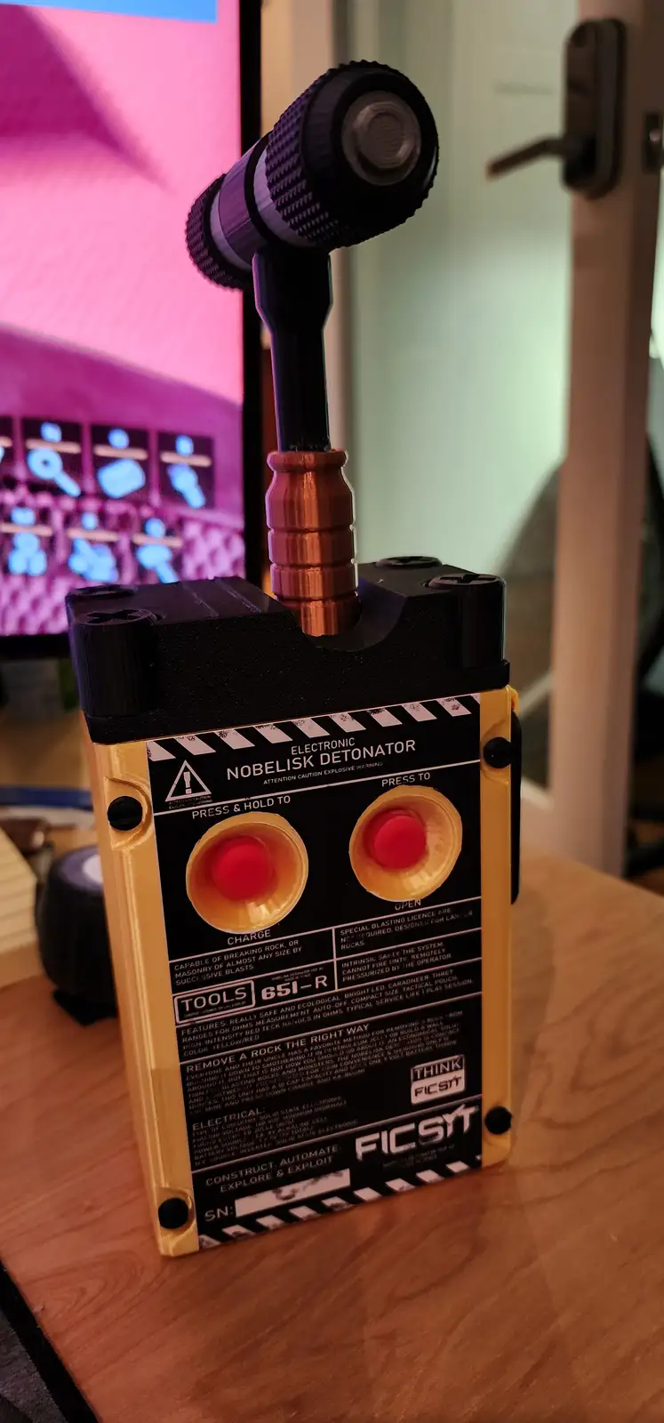 A 3D-printed replica of an 'Electronic Nobelisk Detonator' sits on a wooden desk, featuring two red buttons labeled 'CHARGE'…
