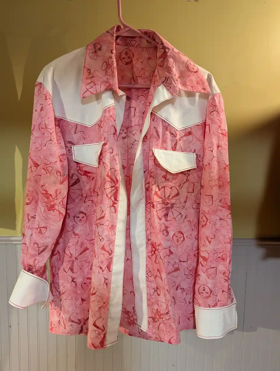 A pink and white western-style shirt with a floral and abstract pattern hangs on a pink hanger against a yellow wall. The…