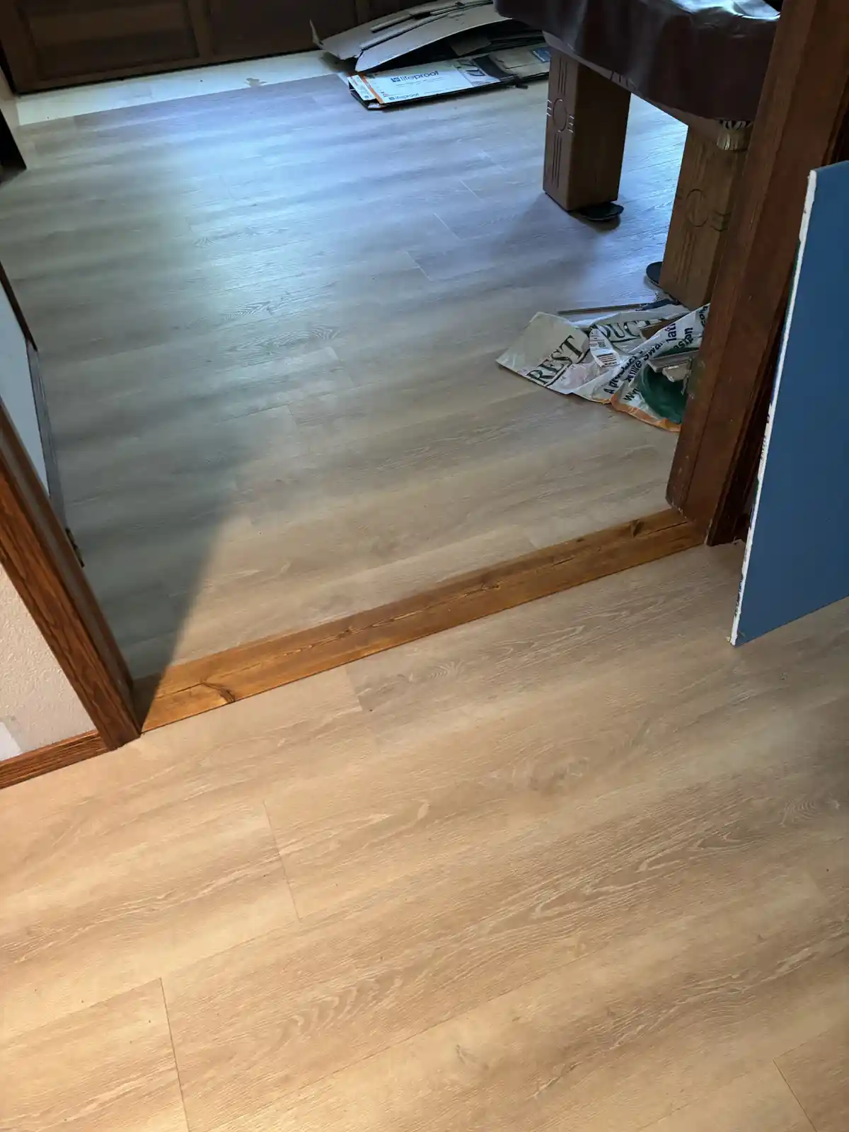 A photo shows a floor with two different shades of wood laminate: a lighter, natural-toned floor in the foreground and a…