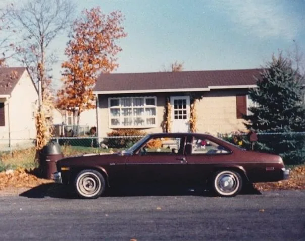 A vintage maroon two-door coupe is parked in front of a suburban house with autumn trees and fallen leaves. The car has…