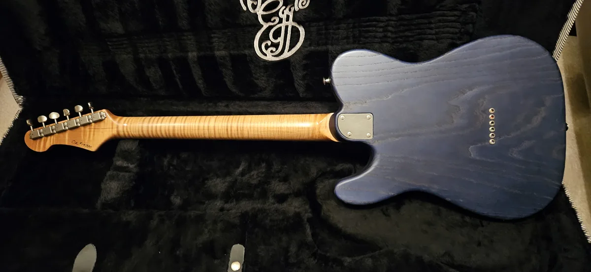 A blue electric guitar with a light wood neck and headstock labeled 'Oz #1450' rests inside a black plush case. The guitar's…