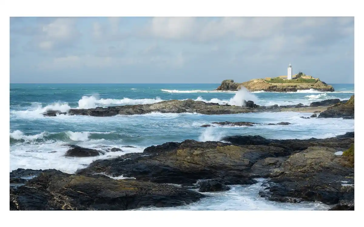 A scenic photograph shows turbulent ocean waves crashing against dark, jagged rocks in the foreground, with a white…