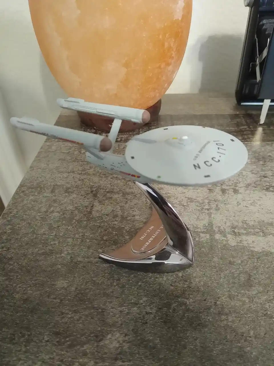 A model of the Star Trek USS Enterprise NCC-1701 sits on a chrome stand on a speckled countertop, with a large orange…
