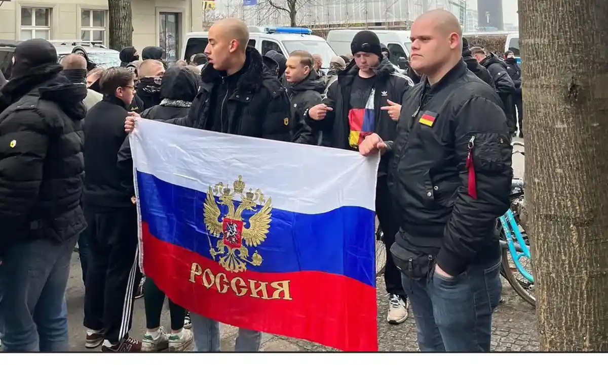 Two men hold a Russian flag with the word 'Россия' written on it, while a third man in a black jacket with a German flag…