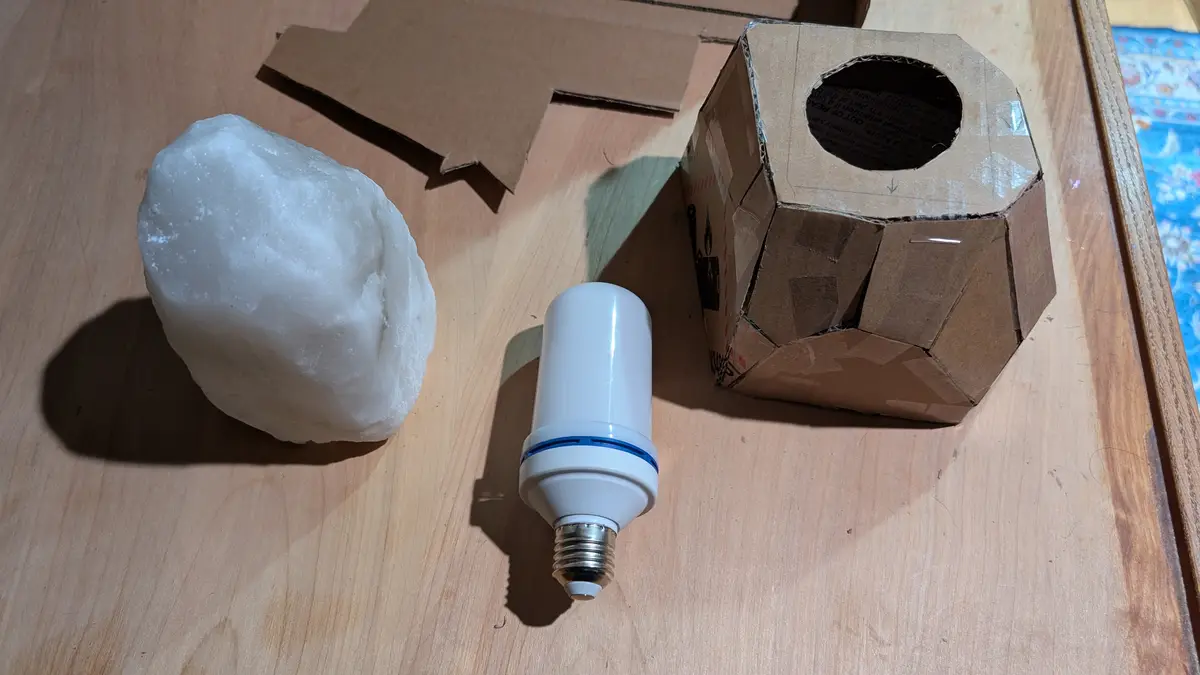 A white, rough-textured salt rock sits next to a standard white LED lightbulb and a handmade cardboard box with a circular…