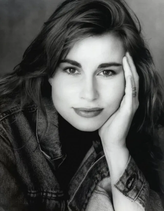 A black-and-white portrait of a woman with wavy hair, wearing a denim jacket, resting her head on her hand with a neutral…