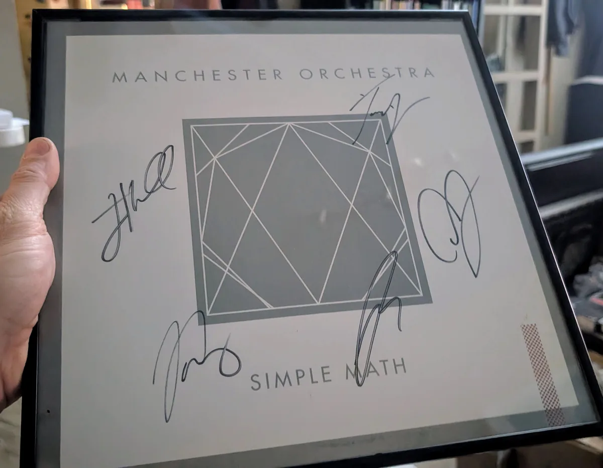 A hand holds a framed album cover for 'Simple Math' by Manchester Orchestra, which has four handwritten signatures overlaid…
