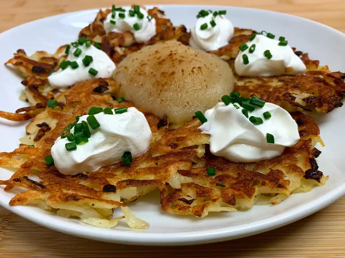 A close-up photo of a white plate holding golden-brown, crispy potato latkes arranged around a single poached pear, all…