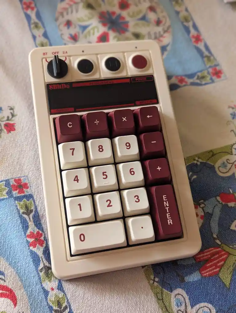 A beige 8BitDo Retro Numpad with maroon and cream-colored keys sits on a floral-patterned fabric. The device features a…