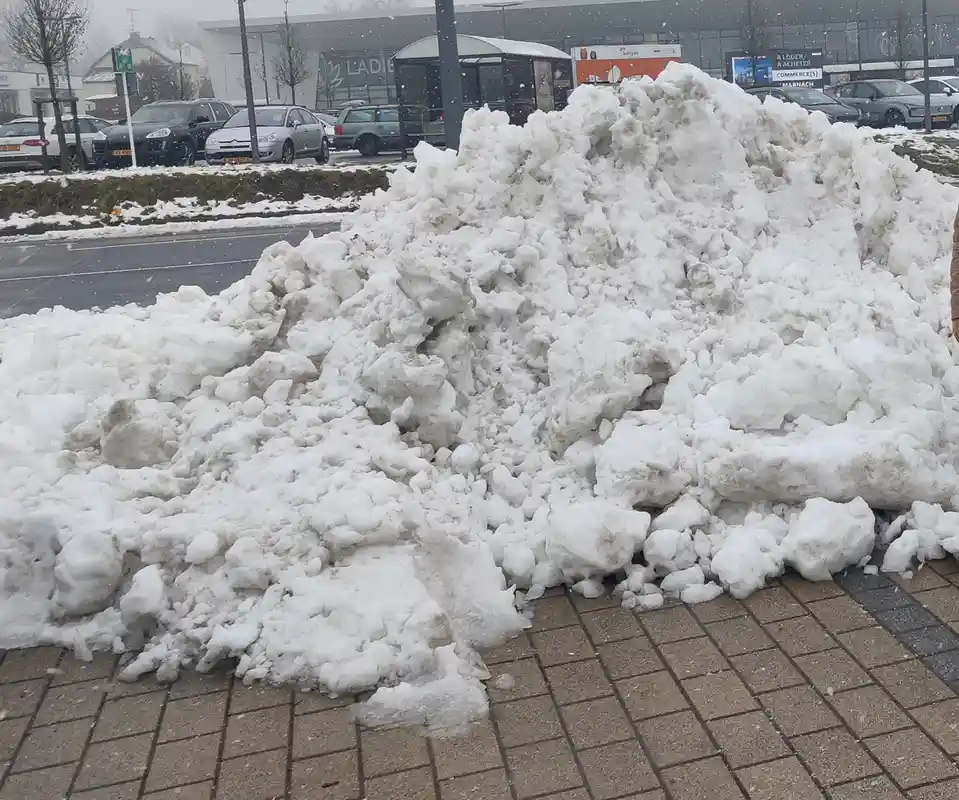 A large, messy pile of snow sits on a paved sidewalk in a snowy urban area, with cars and buildings visible in the…