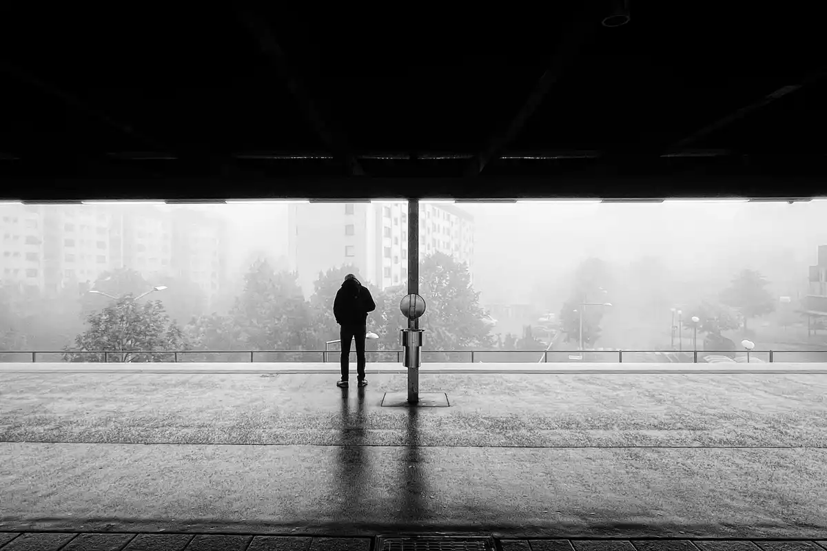A black and white photo shows a person standing alone under an overpass or bridge, looking out at a foggy, urban landscape…