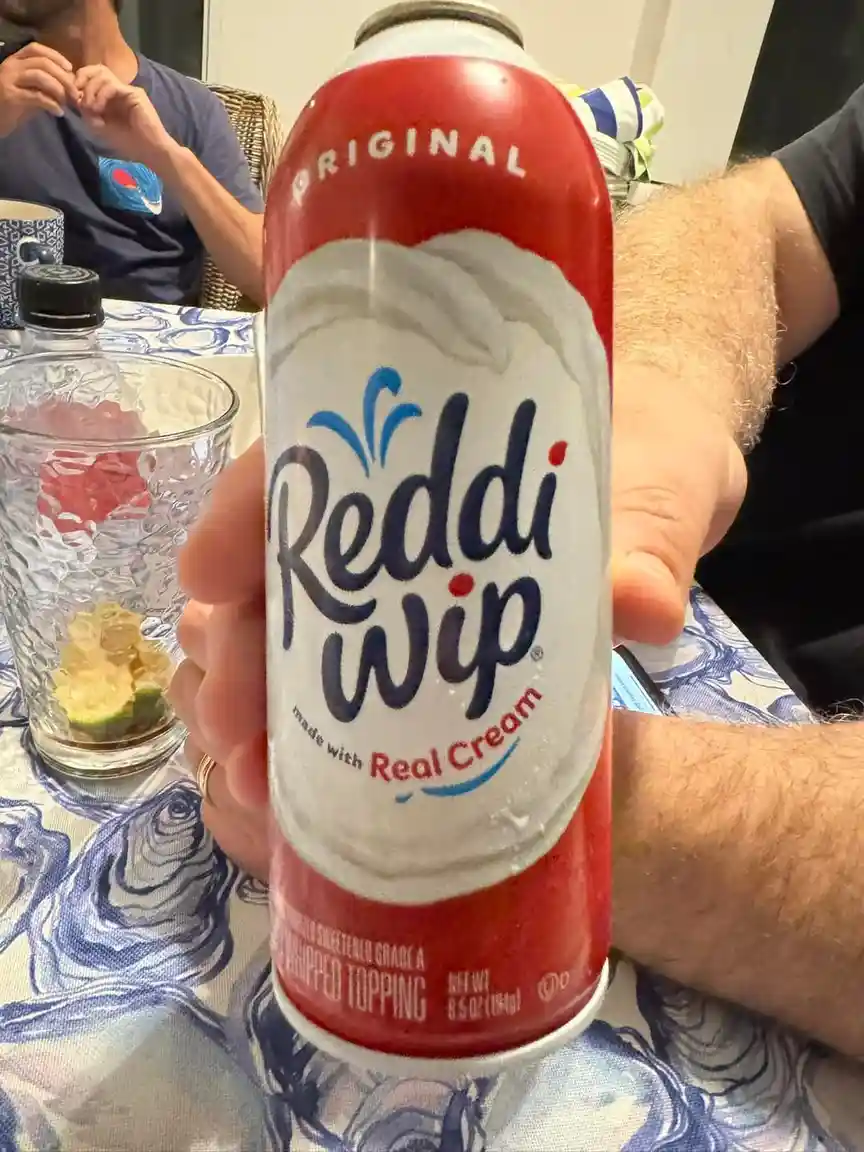 A hand holds a red and white can of Reddi-wip whipped cream, labeled 'Original' and 'made with Real Cream', in front of a…
