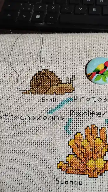 A close-up of a cross-stitch project on light-colored fabric showing a pixelated brown snail labeled 'Snail' and a pixelated…