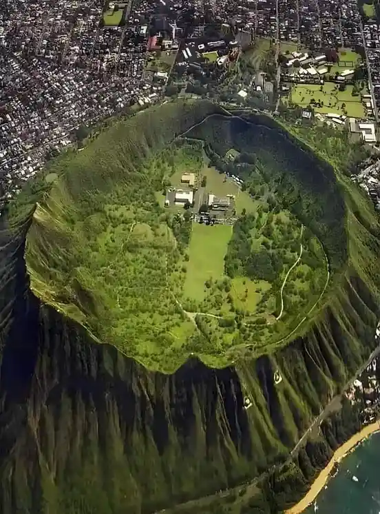 An aerial view of a large, green volcanic crater with buildings and paths inside, surrounded by a dense urban area and…