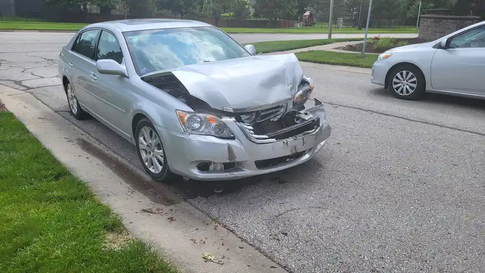 A silver sedan is parked on a residential street with severe front-end damage, including a crumpled hood, broken bumper, and…