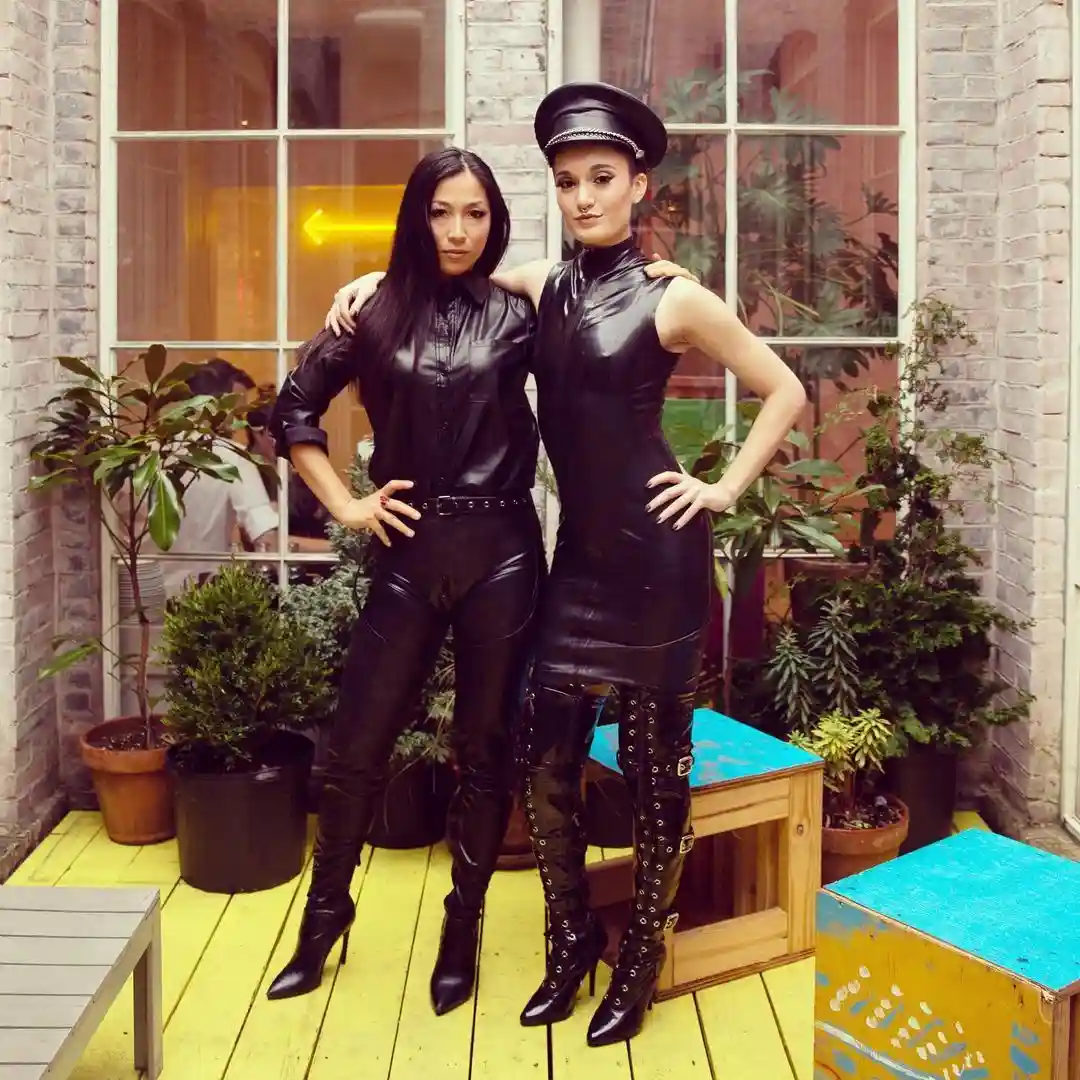 Two women in black leather outfits pose confidently on a yellow wooden deck surrounded by potted plants and brick walls. One…
