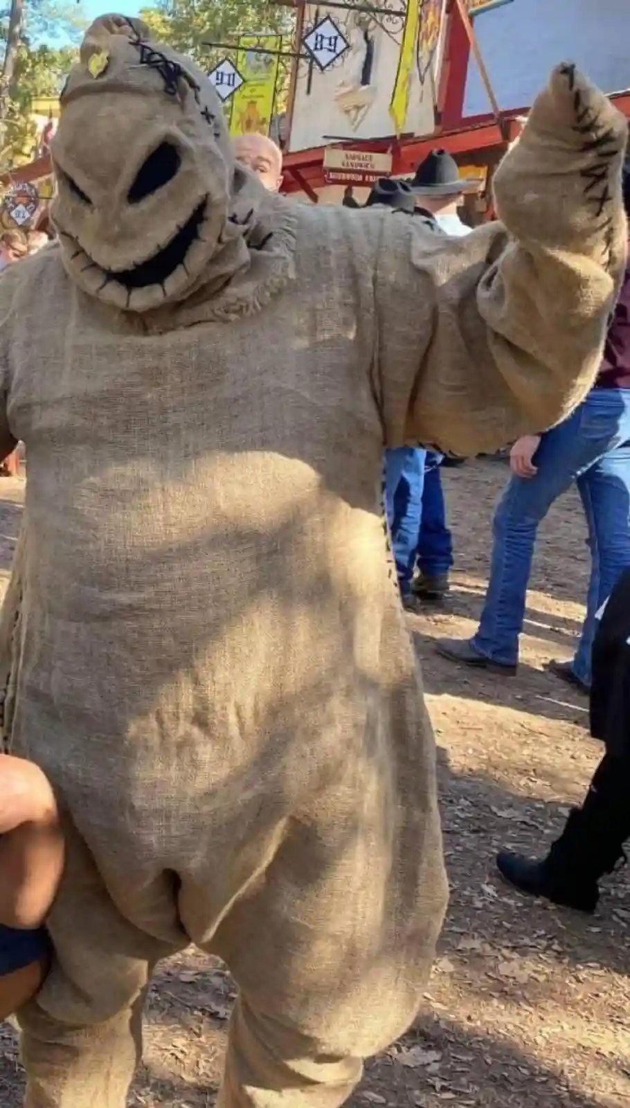 A person in a full-body burlap sack costume resembling a scarecrow with stitched facial features and a wide grin stands…