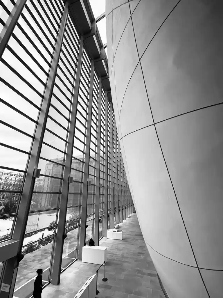 A black-and-white photo shows a long, modern hallway with a curved, segmented wall on the right and tall glass windows on…