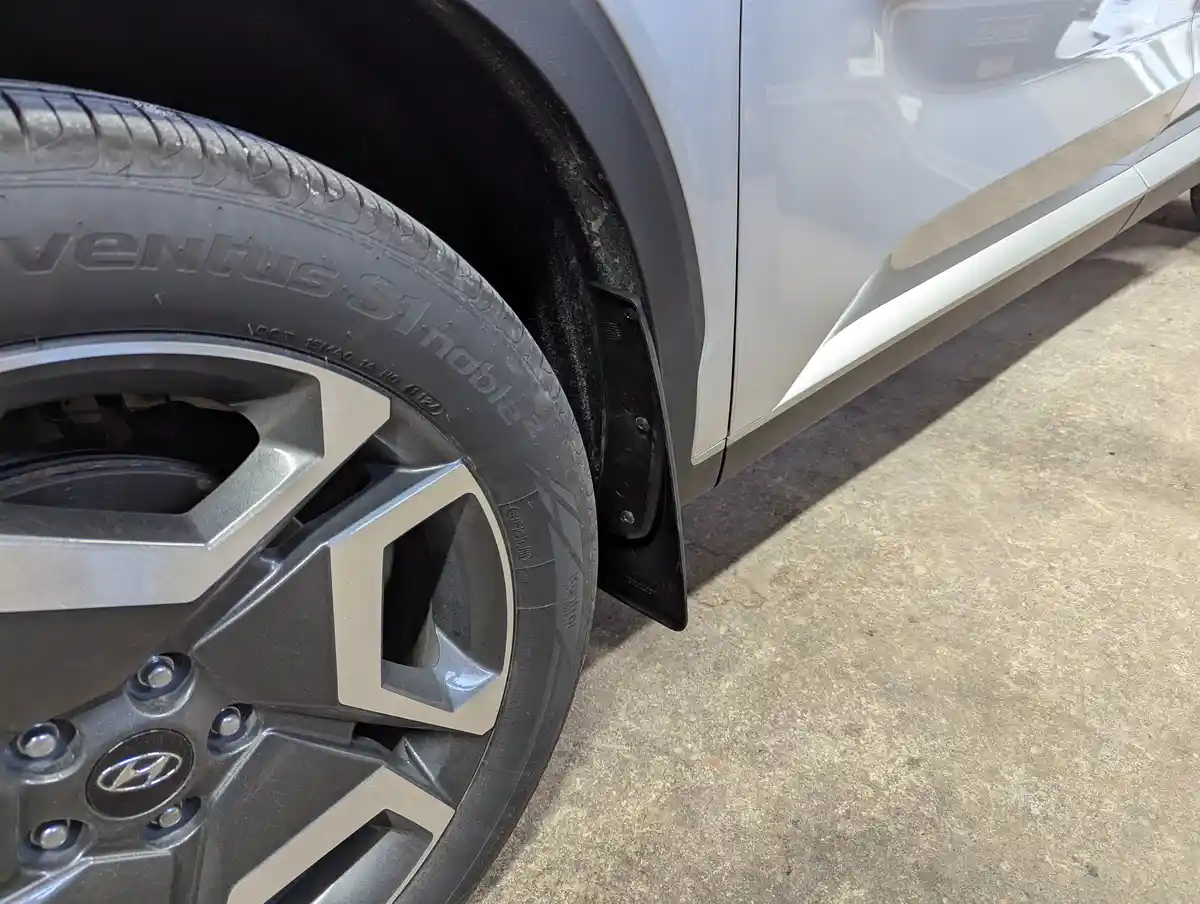 A close-up photo of a silver Hyundai vehicle's front wheel and fender, showing a black mud flap attached to the fender. The…