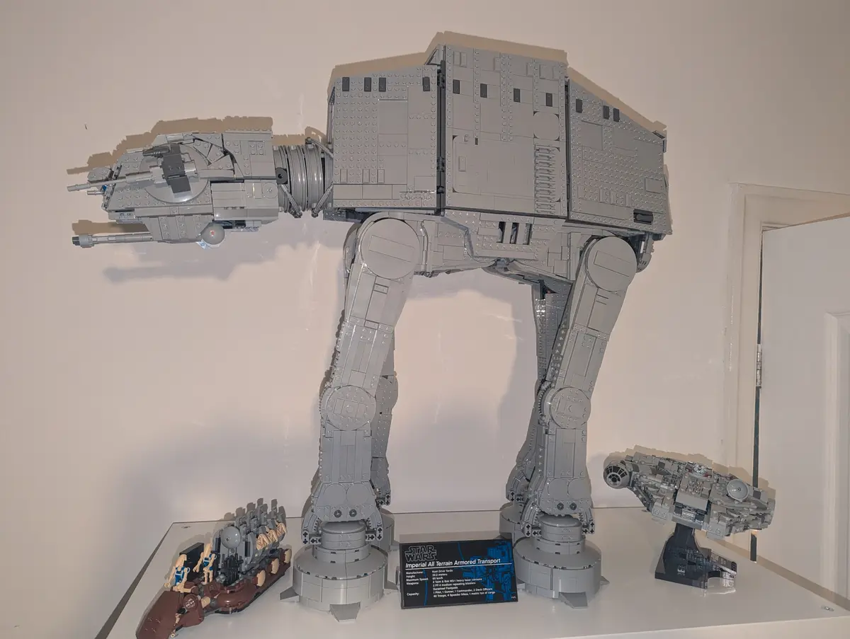 A large, gray LEGO model of an AT-AT walker from Star Wars stands prominently on a white display shelf, flanked by smaller…