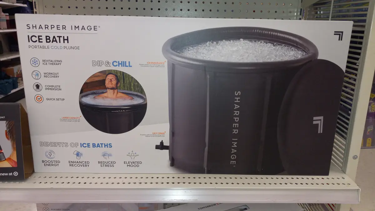 A product box for a Sharper Image portable ice bath is displayed on a retail shelf, featuring an image of a man relaxing in…