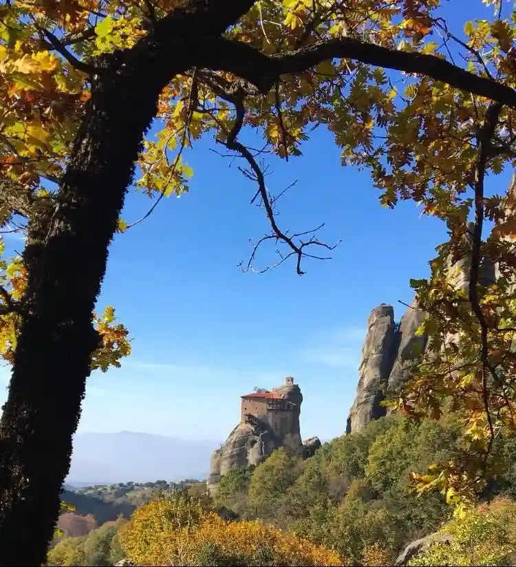 A scenic autumn photo shows a monastery perched atop a rock formation, framed by tree branches with yellow and green leaves…